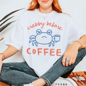 May include: White t-shirt with a crab graphic and the text "crabby before coffee" in coral. The crab is blue and holds a coffee cup. The shirt is a casual, oversized fit.