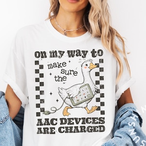 以下が含まれることがあります： タブレットを持った漫画のガチョウのグラフィックと「ON MY WAY TO MAKE SURE THE AAC DEVICES ARE CHARGED」というテキストがプリントされた白いTシャツ。ガチョウは充電ケーブルを繋いで歩いています。