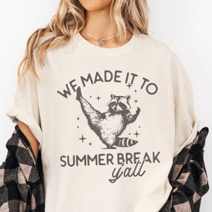 May include: A beige t-shirt with a graphic of a raccoon doing a leg split. The text "We made it to summer break y'all" is printed on the shirt.