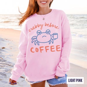 以下が含まれることがあります： オレンジ色の「crabby before coffee」の文字と、コーヒーカップを持った青いカニのグラフィックが特徴の、ライトピンクのスウェットシャツ。クルーネックのスタイルです。