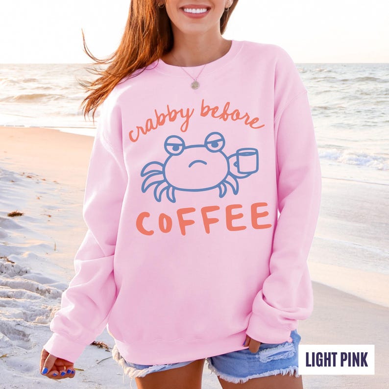以下が含まれることがあります： オレンジ色の「crabby before coffee」の文字と、コーヒーカップを持った青いカニのグラフィックが特徴の、ライトピンクのスウェットシャツ。クルーネックのスタイルです。