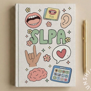 May include: A cream-colored notebook with colorful illustrations and the letters "SLPA". The notebook features drawings of a mouth, ear, hand, brain, speech bubble, and a tablet. A gold pen rests beside the notebook.