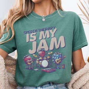 May include: Green t-shirt with a graphic of fruit playing instruments. The text on the shirt reads "Speech Therapy is My Jam".