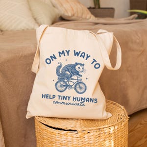 Puede incluir: Una bolsa de lona blanca con texto azul y un gráfico de una zarigüeya montando una bicicleta. El texto dice "On my way to help tiny humans communicate".