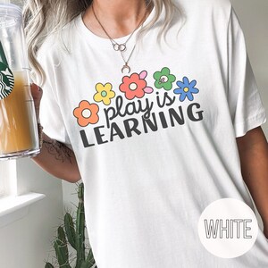 Woman wearing t-shirt that says "Play is Learning" and has cute retro flowers on it for preschool teacher, play therapist, or speech therapist. Gift for teachers or therapists.