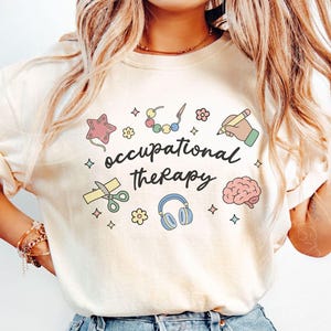 May include: A cream-coloured t-shirt with the words "occupational therapy" in a handwritten font. The shirt features colourful illustrations of a star, scissors, a brain, headphones, and other therapy-related icons.