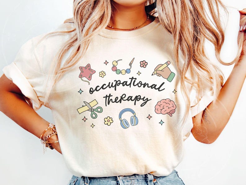 May include: A cream-coloured t-shirt with the words "occupational therapy" in a handwritten font. The shirt features colourful illustrations of a star, scissors, a brain, headphones, and other therapy-related icons.