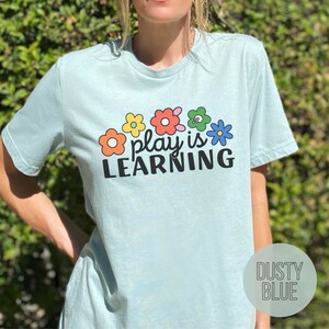 Woman wearing t-shirt that says "Play is Learning" and has cute retro flowers on it for preschool teacher, play therapist, or speech therapist. Gift for teachers or therapists.