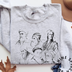 May include: Light gray sweatshirt featuring black line drawings of historical figures, including Pythagoras, Archimedes, and Newton. The names of the figures are written below their portraits. The sweatshirt has a crew neck and long sleeves.
