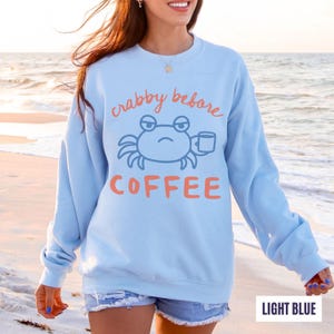 以下が含まれることがあります： 「crabby before COFFEE」の文字と、コーヒーカップを持ったカニのイラストが描かれた水色のスウェットシャツ。 文字とカニはコーラルとブルー。 スウェットシャツはクルーネックです。