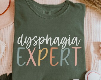 Può includere: T-shirt verde oliva con la scritta "dysphagia EXPERT" in lettere bianche, rosa, gialle e blu. La maglietta è realizzata in materiale morbido e ha uno scollo tondo. Scarpe da ginnastica bianche in tela visibili in basso a destra.