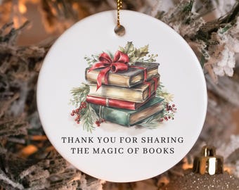 Puede incluir: Adorno navideño de cerámica blanca con lazo dorado para colgar. Presenta una pila de libros adornada con un lazo rojo y vegetación, con el texto "THANK YOU FOR SHARING THE MAGIC OF BOOKS."