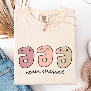 May include: A light beige t-shirt with a graphic of three cartoon faces in pink, peach, and brown. The faces are smiling and the text "never stressed" is printed below the faces.