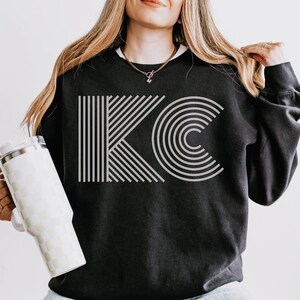 Pu&ograve; includere: Felpa nera con le lettere "KC" in un design geometrico grigio chiaro. La "K" &egrave; composta da linee verticali, e la "C" da archi concentrici. La felpa &egrave; abbinata a jeans chiari e un bicchiere a scacchi.