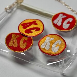 May include: A clear plastic purse with a gold chain strap. The purse has four red and yellow embroidered patches that say "KC" in a retro font.