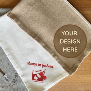 May include: Three folded kitchen towels with a herringbone pattern. One is grey with a shot glass design and the words "shots, shots, shots." Another is white with a cocktail design and the words "always in fashion." The third is beige.