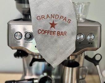 May include: A silver espresso machine with a gray and white striped tea towel draped over the top. The tea towel has the text "Grandpas Coffee Bar" embroidered on it in brown thread with a red star.