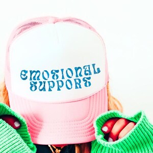 Puede incluir: Gorra de camionero rosa y blanca con el texto azul "EMOTIONAL SUPPORTS". La gorra se usa con un suéter verde.