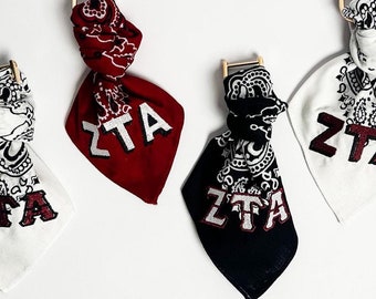 Puede incluir: Cuatro bandanas con las letras "ZTA" en blanco sobre un fondo rojo, negro y blanco. Las bandanas están atadas y colgando de una varilla de madera.