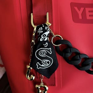 May include: A red bag with a gold-colored chain and a black chain. A black and white bandana is attached to a gold-colored hook. A black bag with a white embroidered "S" is also attached.
