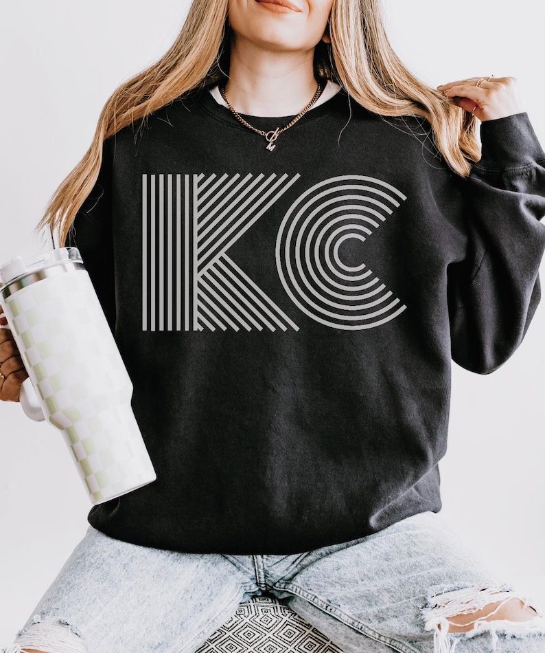 Pu&ograve; includere: Felpa nera con le lettere "KC" in un design geometrico grigio chiaro. La "K" &egrave; composta da linee verticali, e la "C" da archi concentrici. La felpa &egrave; abbinata a jeans chiari e un bicchiere a scacchi.