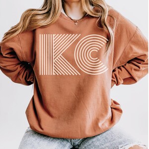 Pu&ograve; includere: Una felpa color ruggine con le lettere "KC" in un design geometrico color crema. La "K" &egrave; composta da linee verticali e diagonali, mentre la "C" &egrave; formata da archi concentrici. La felpa &egrave; di stile casual.