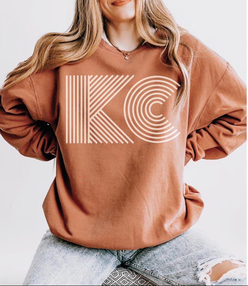 Pu&ograve; includere: Una felpa color ruggine con le lettere "KC" in un design geometrico color crema. La "K" &egrave; composta da linee verticali e diagonali, mentre la "C" &egrave; formata da archi concentrici. La felpa &egrave; di stile casual.