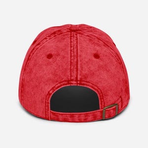 May include: A red baseball cap with a curved brim and a metal buckle closure. The cap has a vintage, worn look and a solid color design. The back of the cap is visible.