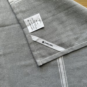 May include: Gray linen kitchen towel with white stripes and a herringbone pattern. The towel has a label that says "Linen + Home" and includes care instructions.