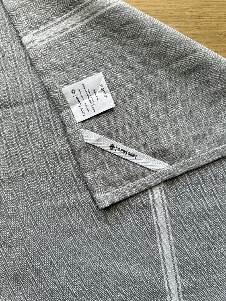 May include: Gray linen kitchen towel with white stripes and a herringbone pattern. The towel has a label that says "Linen + Home" and includes care instructions.