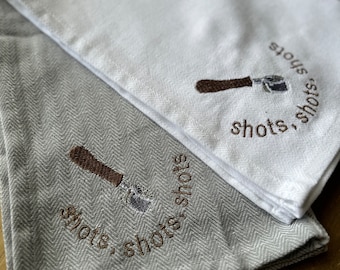Puede incluir: Dos toallas de cocina, una blanca y otra gris con un patrón de espiga. Ambas toallas presentan una imagen bordada de un portafiltro de café y el texto "shots, shots, shots".