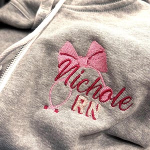 May include: A heather grey zip-up hoodie featuring an embroidered design on the chest. The embroidery includes a pink bow, a stethoscope outline, and the text "Nichole RN" in dark pink script lettering with light pink block letters for RN.
