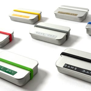 Könnte beinhalten: Sechs kleine silberne Dosen mit bunten Gummibändern drumherum. Jede Dose hat ein weißes Etikett mit der Aufschrift "GAME DAY" in schwarzen Buchstaben.