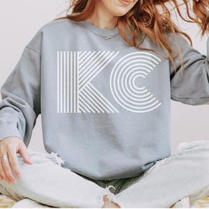 Pu&ograve; includere: Una felpa blu chiaro con le lettere bianche "KC" in un design geometrico audace. La "K" &egrave; composta da linee verticali e diagonali, mentre la "C" &egrave; formata da semicerchi concentrici. La felpa &egrave; uno stile casual e confortevole.