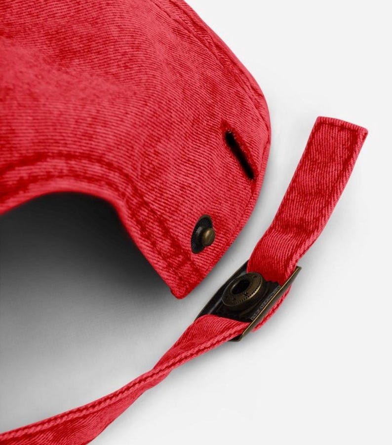 May include: Close-up of a red baseball cap with a curved brim. The cap features a metal clasp and a fabric strap for adjustment. The cap is made of a textured fabric.