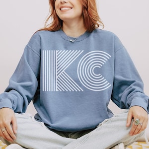 Pu&ograve; includere: Una felpa blu chiaro con le lettere bianche "KC" in un design geometrico. La "K" &egrave; composta da linee verticali e la "C" &egrave; composta da archi concentrici. La felpa ha maniche lunghe e girocollo.