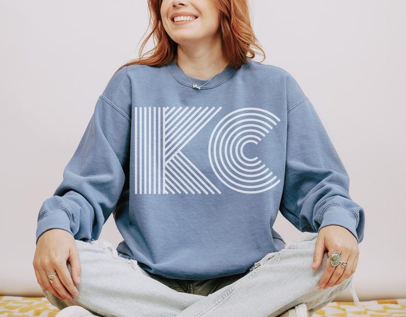 Pu&ograve; includere: Una felpa blu chiaro con le lettere bianche "KC" in un design geometrico. La "K" &egrave; composta da linee verticali e la "C" &egrave; composta da archi concentrici. La felpa ha maniche lunghe e girocollo.
