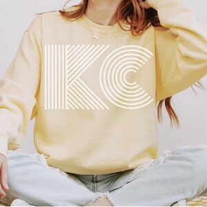 Pu&ograve; includere: Una felpa giallo pallido con le lettere bianche "KC" stampate sul davanti. La "K" &egrave; composta da linee verticali e la "C" da archi concentrici. La felpa ha maniche lunghe e girocollo.