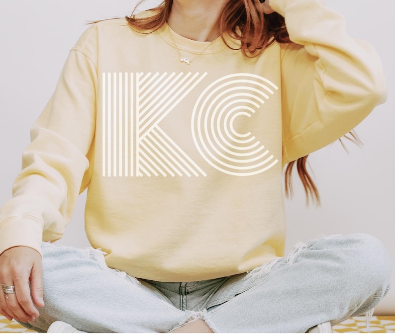 Pu&ograve; includere: Una felpa giallo pallido con le lettere bianche "KC" stampate sul davanti. La "K" &egrave; composta da linee verticali e la "C" da archi concentrici. La felpa ha maniche lunghe e girocollo.