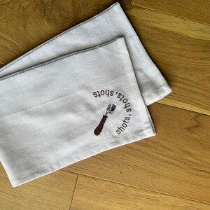 May include: White kitchen towel with brown embroidery of a coffee tamper and the text "shots, shots, shots".