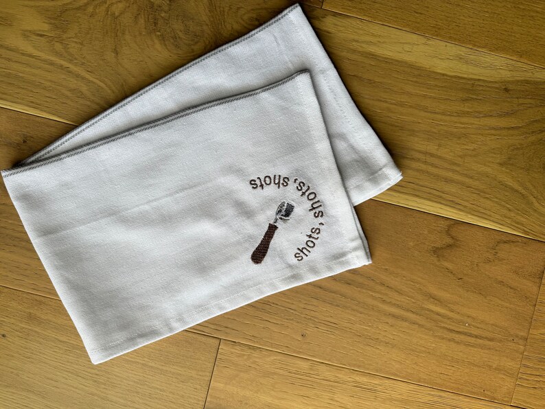 May include: White kitchen towel with brown embroidery of a coffee tamper and the text "shots, shots, shots".