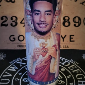 Op de afbeelding: Een lange glazen kaars met een portret van een man in religieuze beelden. De kaars heeft een blauwe band met kruisen en een wit wasinterieur. De achtergrond omvat een Ouija-bord en een zwarte mat met symbolen.