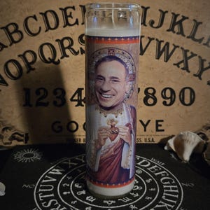 May include: A tall glass candle with a printed image of a smiling man in religious attire, holding a heart. The candle has a white wax interior and is set against a Ouija board background.