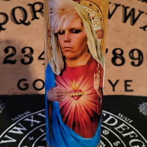 May include: A glass candle with a white wax interior and a colorful image of a person with blonde hair wearing a blue and red robe. The image has a heart with flames and gold teeth. The candle is sitting on a black surface with a white circle and the words "YES" and "LMNOPQR" printed on it.