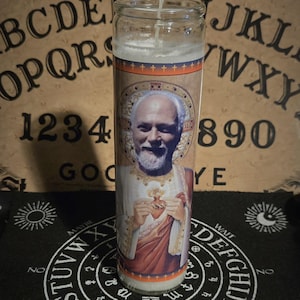 May include: A glass candle featuring a portrait of a man in religious clothing. The candle has a white wax top and a label with an image of a man with a white beard. The candle is on a black surface with white text and symbols.