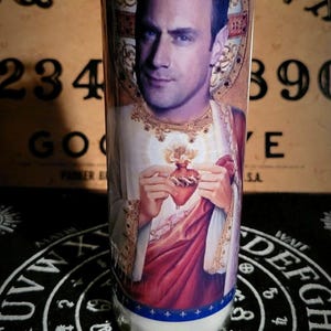 Könnte beinhalten: Ein Glas-Kerzengefäß mit einer weißen Kerze darin. Das Gefäß hat ein gedrucktes Bild eines Mannes mit einem Herzen in seinen Händen. Das Bild ist von einem goldenen Rahmen umgeben. Das Gefäß steht auf einer schwarzen Oberfläche mit einem weißen Kreis und dem Wort "YES" in der Mitte.