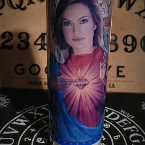 Puede incluir: Una vela de vidrio transparente con una imagen impresa de una mujer en una representación religiosa roja y azul. La vela está sobre una superficie negra con letras y símbolos blancos. Un tablero Ouija está en el fondo.