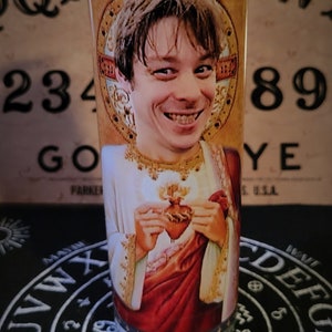 Może przedstawiać: Szklana świeca z białą świecą w środku. Świeca ma nadrukowany obraz mężczyzny trzymającego serce w dłoniach. Obraz jest otoczony złotą ramą. Świeca stoi na czarnej powierzchni z białym kołem z literami i symbolami.