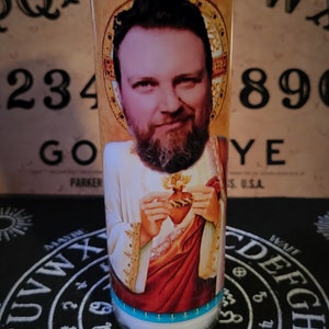 Może przedstawiać: Wysoka, przezroczysta szklana świeca z portretem mężczyzny w stroju religijnym. Świeca ma białe wnętrze z wosku oraz niebieską i złotą etykietę. Tło obejmuje tablicę Ouija i czarną matę z białymi literami.