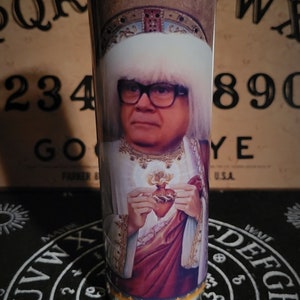 May include: A glass candle with a white wax base and a colorful image of a man wearing a white fur hat and glasses, holding a heart. The candle is sitting on a black surface with a white circle design.